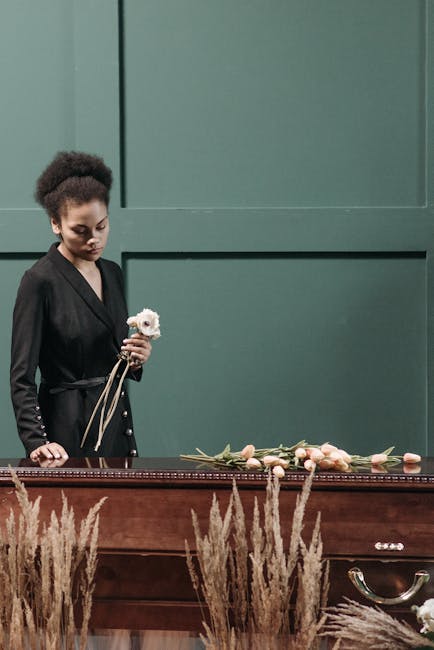 A woman with dark skin and curly hair styled in a high puff stands beside a wooden casket, holding a single white flower. She is dressed in a black, long-sleeved outfit. On the casket, there are sever