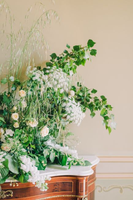 An elegant floral arrangement placed on a polished wooden table with a white marble top, featuring a mix of greenery and delicate flowers. The greenery includes lush, broad leaves and trailing ivy, co