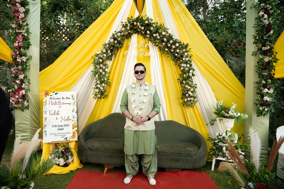 A group of well-dressed wedding guests gathered outside a grand, historic building with classical columns and large windows, celebrating a wedding ceremony. The setting features vibrant floral arrange