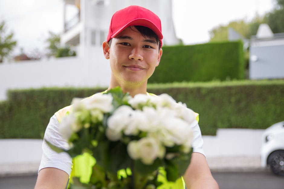 A delivery person wearing a red cap and a high-visibility vest is holding out a bouquet of fresh white roses and greenery towards the camera, with a friendly expression. The bouquet is neatly arranged