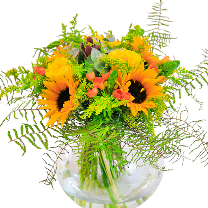 With its sun-drenched palette and abundance of texture, this bouquet from Bexley Florist exudes a sense of joy and togetherness. The centrepiece sunflowers boast bold, golden petals radiating from deep, inviting centres, reminiscent of flower beds brightening the edge of Bexley's Danson Park during the height of summer. Tucked elegantly amidst the sunflowers, the yellow garden roses spiral open in creamy, compact formations; each petal feels tender and full, a nod to the classic garden blooms often seen in local Bexley gardens. Contrasting with the soft petals, clusters of shiny red Hypericum berries dot the composition, their colour as vibrant as the bustling activity found along Bexley's Broadway. Fine, feathery ferns and tapering filler stems create a gentle cascade, introducing movement, depth, and a touch of wildness. Blue-green eucalyptus adds a subtle aromatic hint and softens the arrangement with its cool tones. Contained in a modern, round glass vase, every stem is visible, the clear water enhancing their natural lines and amplifying the bouquet's freshness. This composition shines as a centrepiece for happy moments and meaningful celebrations-a birthday, anniversary, or just a brightening gesture. Crafted with pride and local expertise, each flower and leaf reflects the nurturing hands of Bexley's florists, bringing the uplifting energy of the community directly into your home.